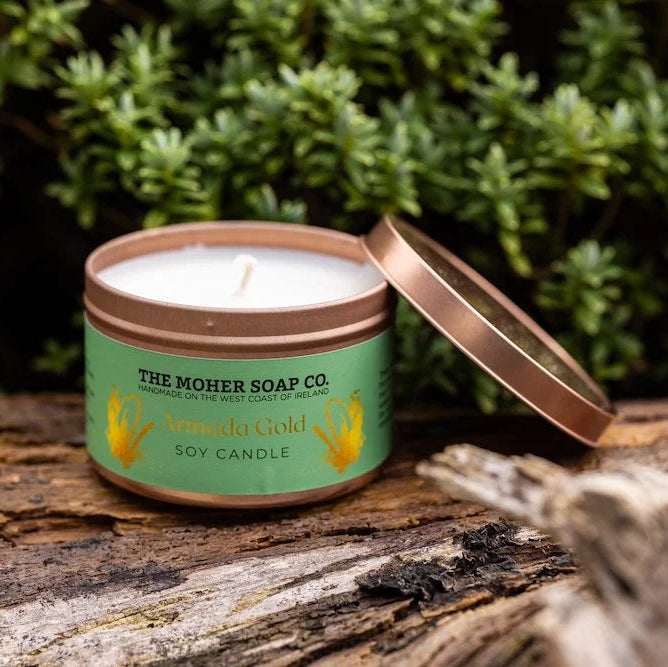Candle in a tin from The Moher Soap Co. on a wooden surface with greenery in the background