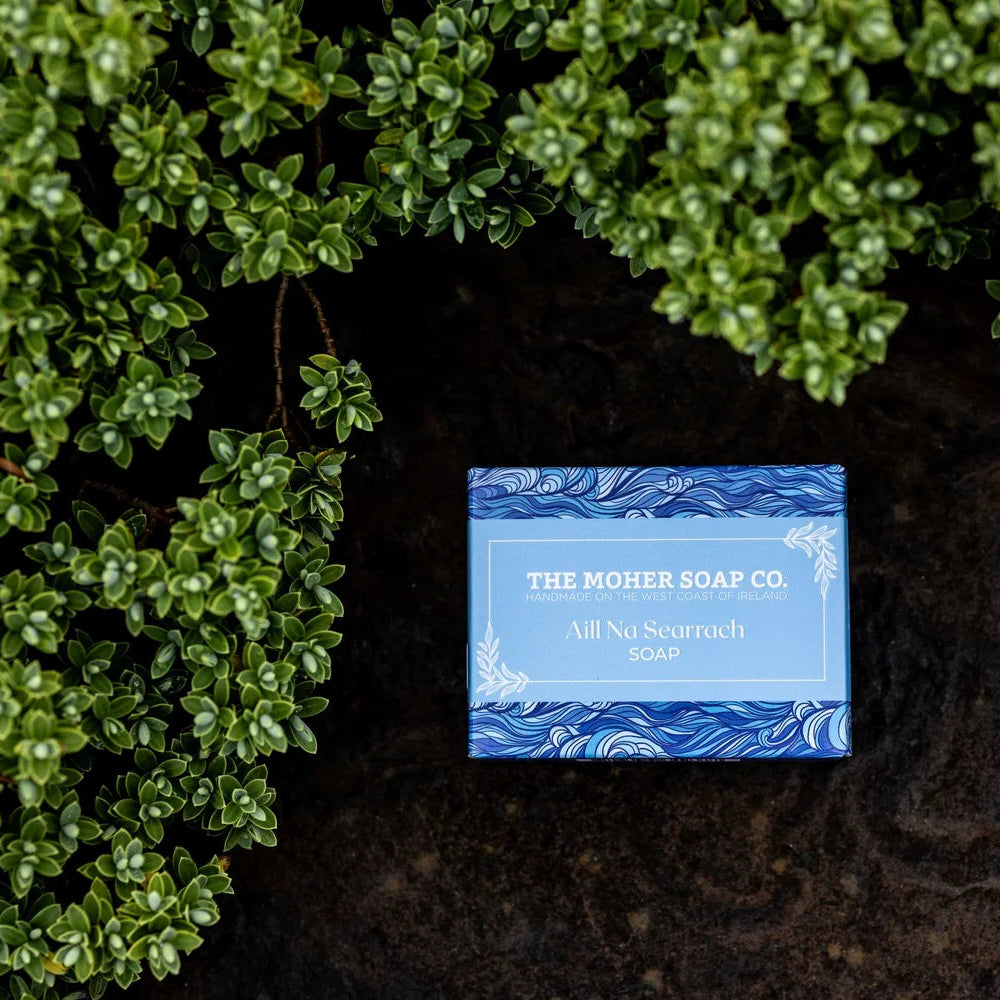 Blue soap bar with 'The Nother Soap Co' label on a dark surface surrounded by green foliage