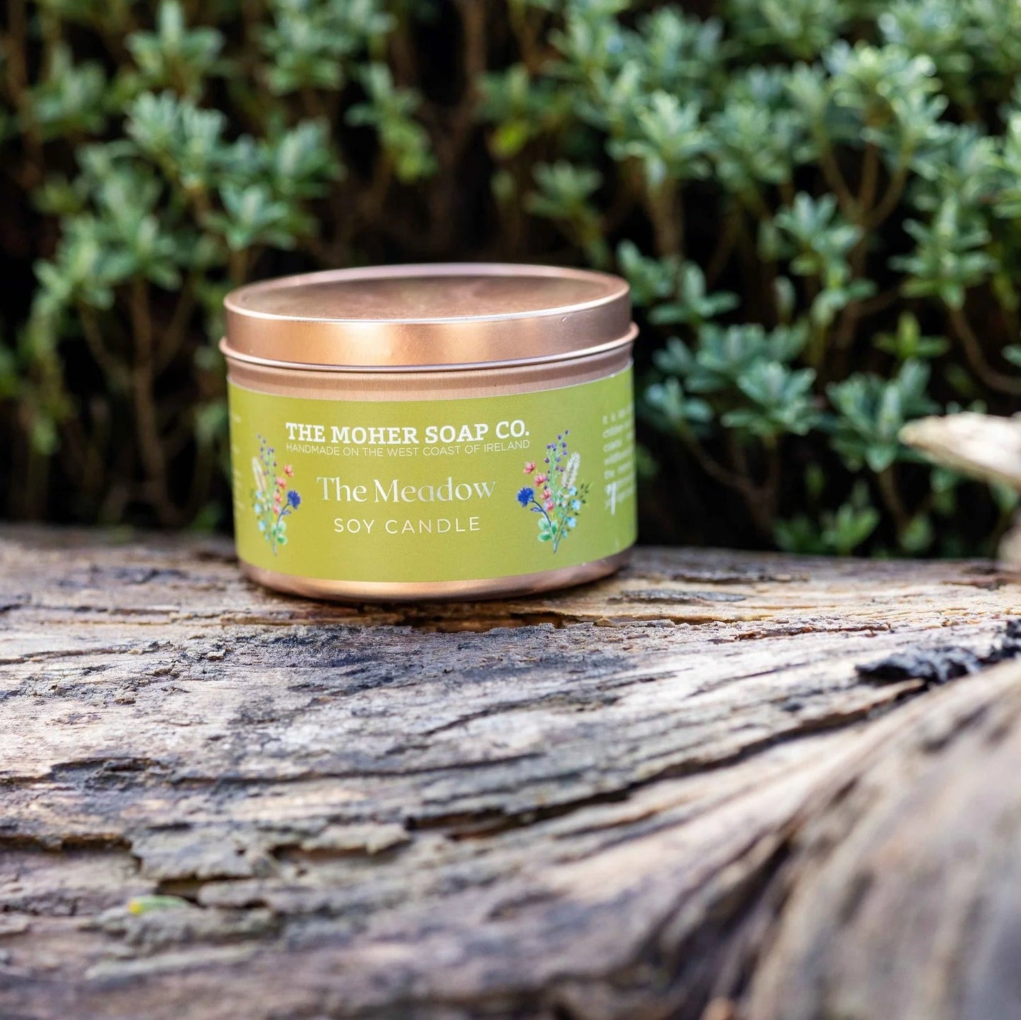 Candle in a tin from The Mother Soap Co. on a wooden surface with greenery in the background