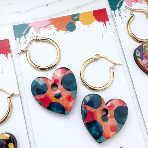 Colorful heart-shaped earrings on branded cards with 'themessybrunette.com' branding.