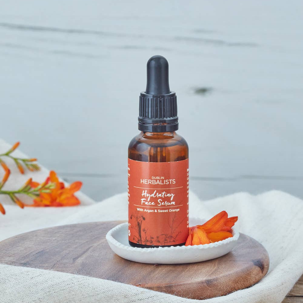 Bottle of Herbalists Hydrating Face Serum on a wooden surface with carrots and flowers.