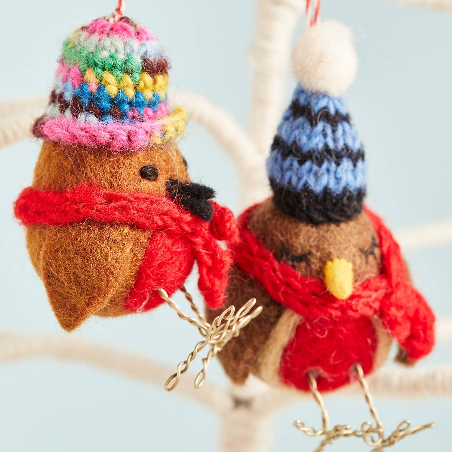 Two hand-knitted felt robins with colorful hats and scarves, hanging from a branch with decorative red berries on the branches and a blue sky in the background.