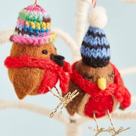 Two hand-knitted felt robins with colorful hats and scarves, hanging from a branch with decorative red berries on the branches and a blue sky in the background.