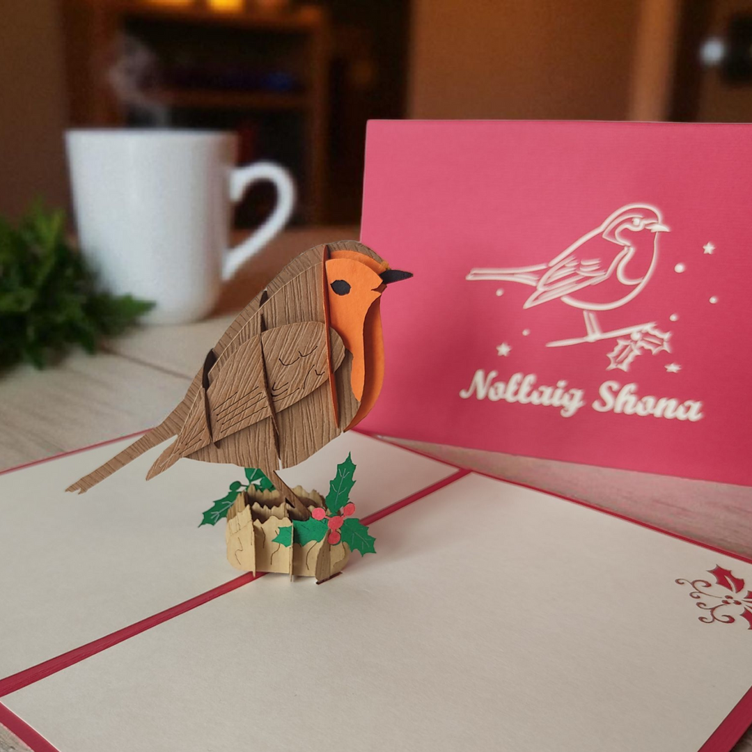 Pop-up card with a robin design and a pink card with 'Nollaig Shona' text on a table.