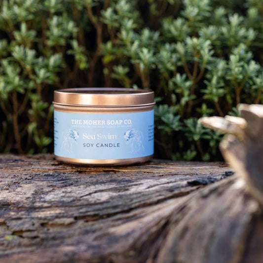 Scented candle in a tin with a blue label on a wooden surface with greenery in the background