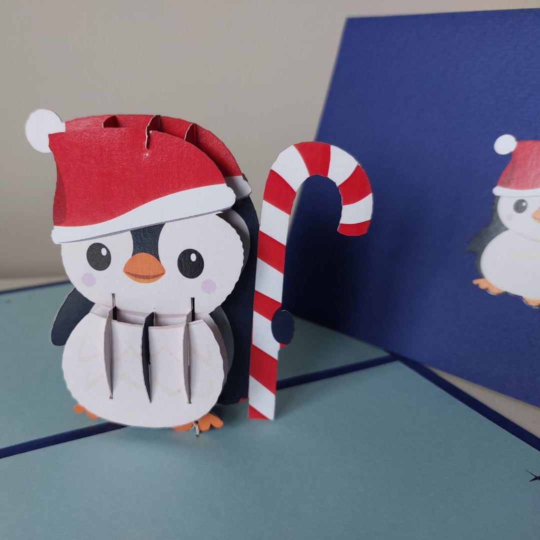 3D paper craft of a penguin with a Santa hat and candy cane on a blue card.