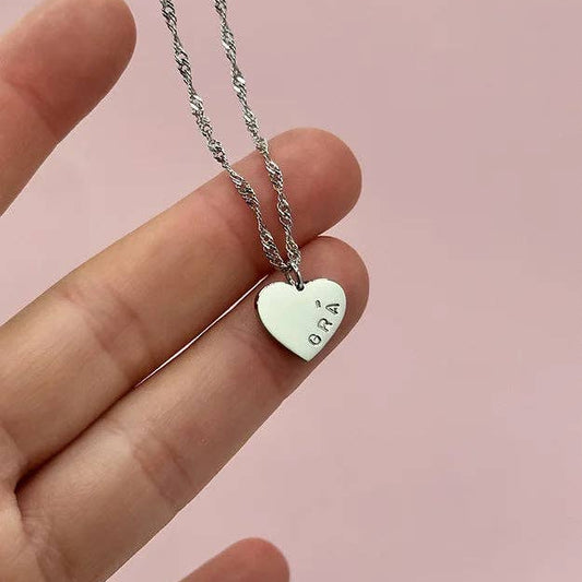 Heart-shaped pendant necklace with 'ORA' engraving held by a hand against a pink background
